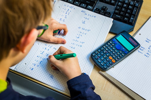 student with graphing calculator sitting at desk writing out equations with laptop in front of them